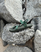 A single men's natural green EXPLOre Roman Sandal on a rugged coastal granite boulder, highlighting its durable minimalist strap design for all-terrain wear.