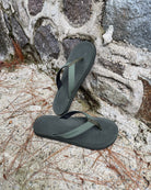 Men's natural green Flow Thong Sandals leaning against a textured stone wall, conveying a casual yet authentic style ready for everyday adventures.