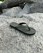 A single men's natural green Flow Thong Sandal resting on rugged coastal rock, showcasing its minimalist design for effortless outdoor presence.