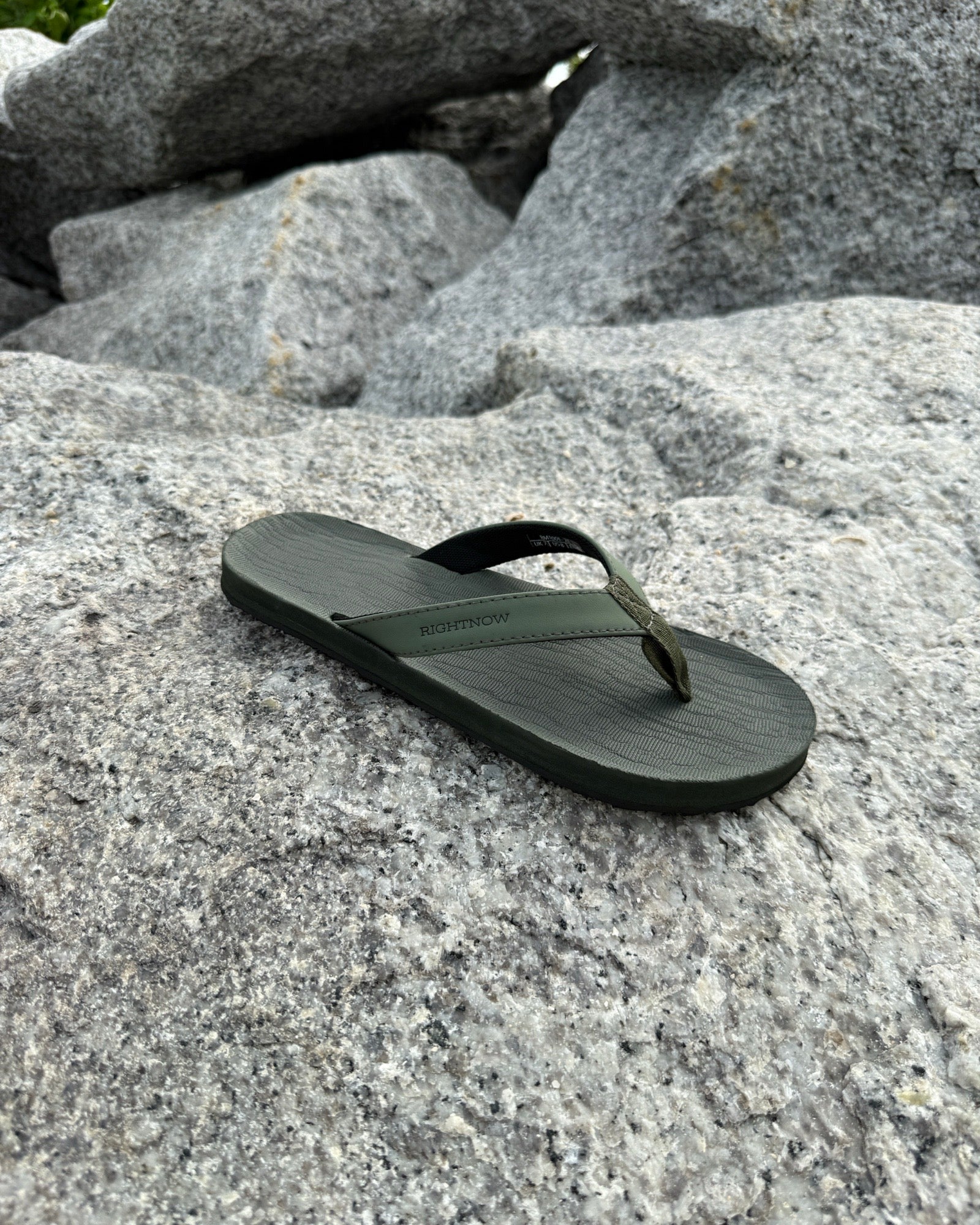 A single men's natural green Flow Thong Sandal resting on rugged coastal rock, showcasing its minimalist design for effortless outdoor presence.