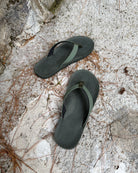 Top-down view of men's forest-tone Flow Thong Sandals on concrete ground with scattered pine needles, highlighting their simple design for natural and urban exploration.