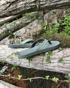 Men's natural green Flow Thong Sandals displayed on a moss-covered fallen log amidst lush greenery, perfect for authentic moments of relaxation at a campground in nature.
