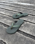 A pair of men's natural green Flow Thong Sandals on a dark, textured concrete surface, showcasing their minimalist comfort for modern living.