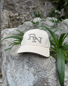 A sand beige Ignite RN logo cap resting on a large, sunlit rock in a quiet garden, embodying a moment of simple, peaceful presence outdoors.