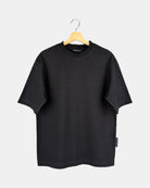 Unisex Moment oversized plain t-shirt in matt black, made from premium cotton, shown from the front to feature its butterfly-knitted crewneck collar on a hanger.