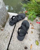 A pair of women's matt black EXPLOre Roman Sandals on a stone ledge by a tranquil stream, showing a simple, authentic lifestyle connected with nature.