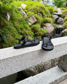 Women's matt black EXPLOre Roman Sandals on a bridge over a serene waterfall and lush greenery, perfect for travel and finding a quiet moment of presence in nature.