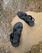 Top-down view of women's elegant-tone EXPLOre Roman Sandals on wet, sandy riverside ground, showing their simple design and flexible straps for any natural terrain.