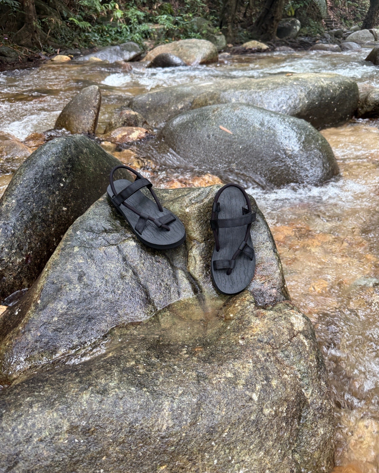 Waterproof women's matt black EXPLOre Roman Sandals on a wet boulder in a flowing jungle stream, highlighting their anti-slip soles for secure footing on any terrain.