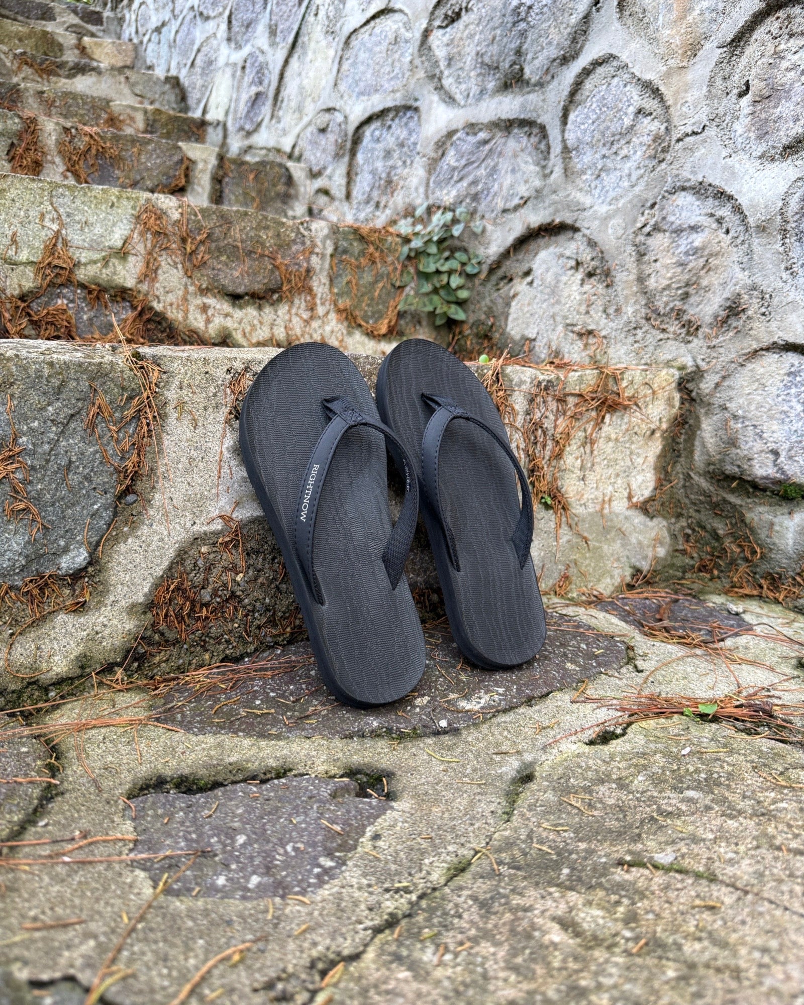 A pair of women's matt black Flow Thong Sandals on rustic stone steps, perfect for a simple and authentic journey through historical or natural paths.