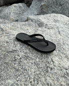 A single women's matt black Flow Thong Sandal resting on rugged coastal rock, showcasing its minimalist design for effortless outdoor presence.