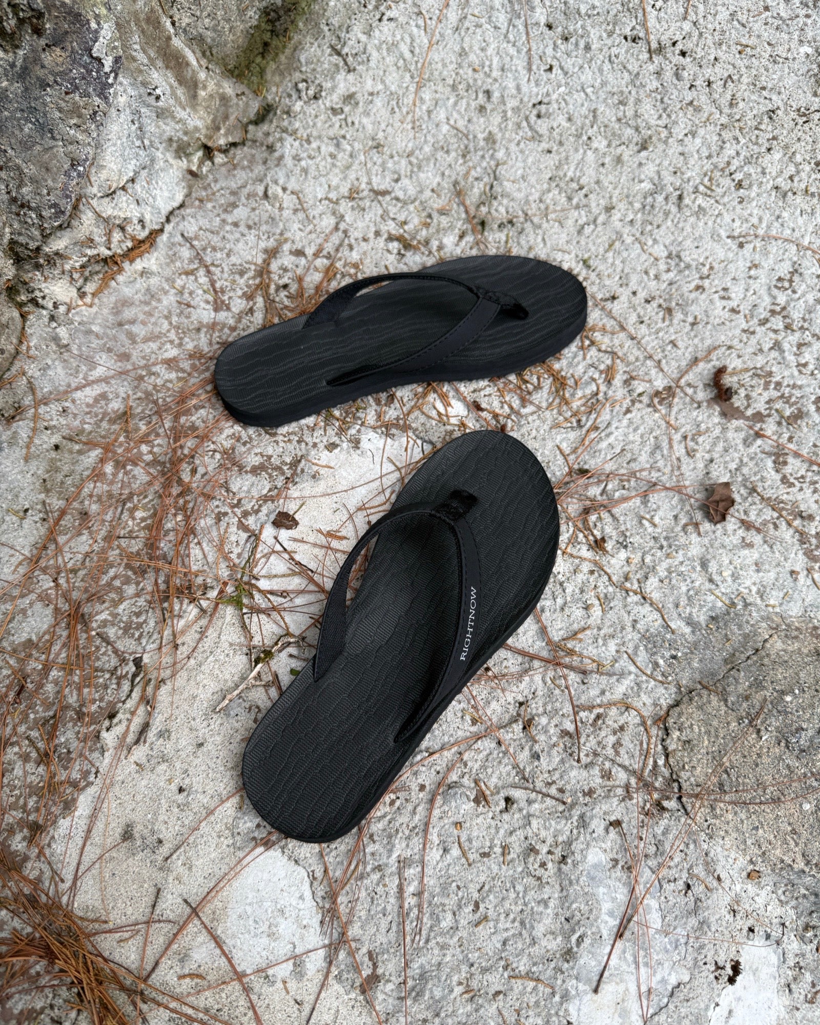 Top-down view of women's elegant-tone Flow Thong Sandals on concrete ground with scattered pine needles, highlighting their simple design for natural and urban exploration.