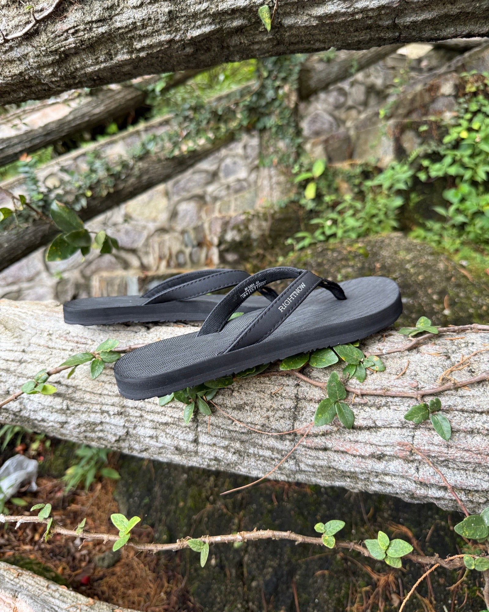 Women's matt black Flow Thong Sandals displayed on a moss-covered fallen log amidst lush greenery, perfect for authentic moments of relaxation at a campground in nature.