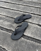 A pair of women's matt black Flow Thong Sandals on a dark, textured concrete surface, showcasing their minimalist comfort for modern living.