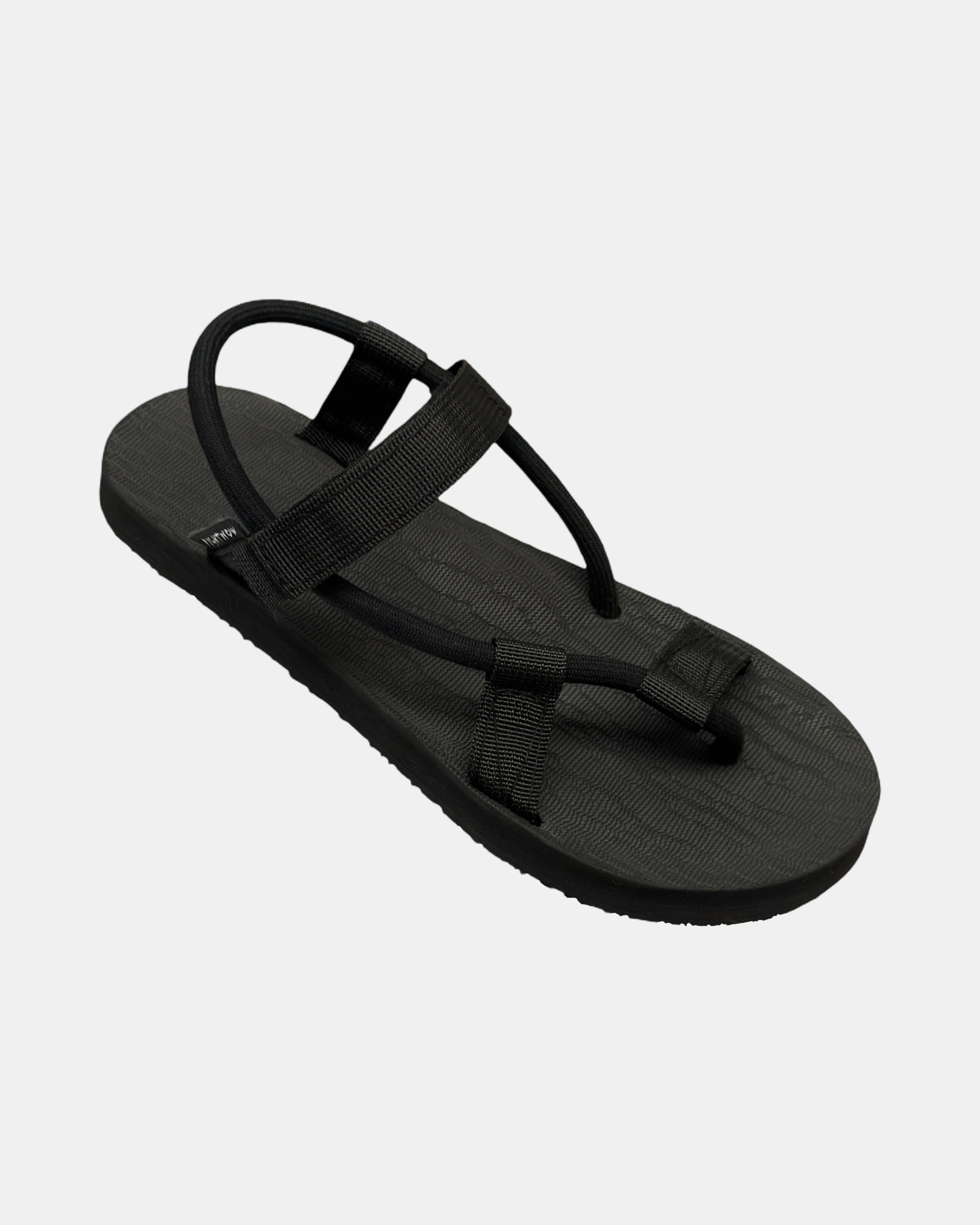 A 45-degree angled view of a women's matt black rubber Roman-style sandal with a single 360° flexible strap around the ankle.