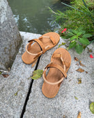A pair of women's earth brown EXPLOre Roman Sandals on a stone ledge by a tranquil stream, showing a simple, authentic lifestyle connected with nature.