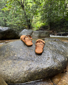 Women's earth brown EXPLOre Roman Sandals on a river rock with a lush tropical rainforest backdrop, the perfect footwear for authentic tropical exploration.