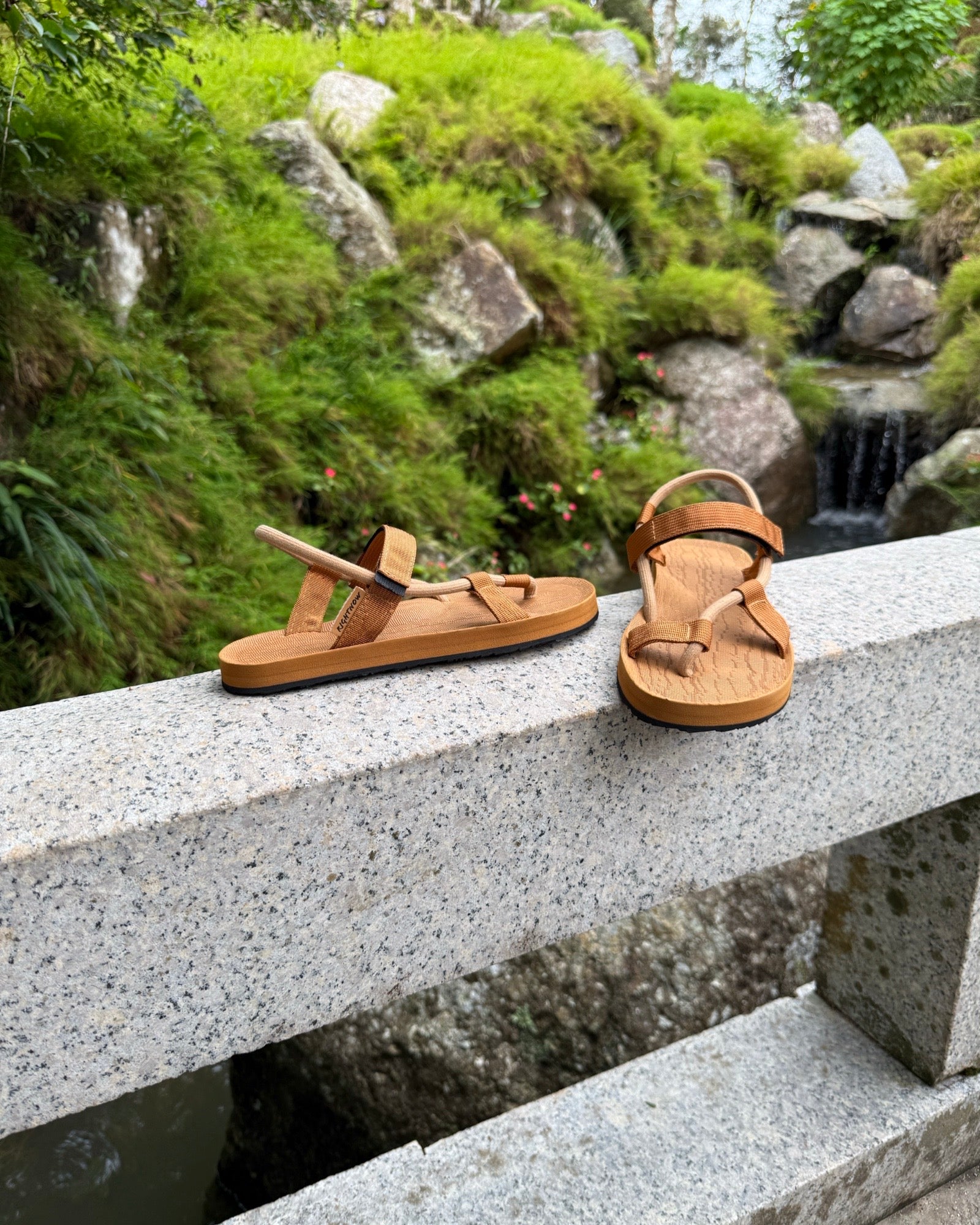 Women's earth brown EXPLOre Roman Sandals on a bridge over a serene waterfall and lush greenery, perfect for travel and finding a quiet moment of presence in nature.