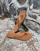 Women's earth brown Flow Thong Sandals leaning against a textured stone wall, conveying a casual yet authentic style ready for everyday adventures.
