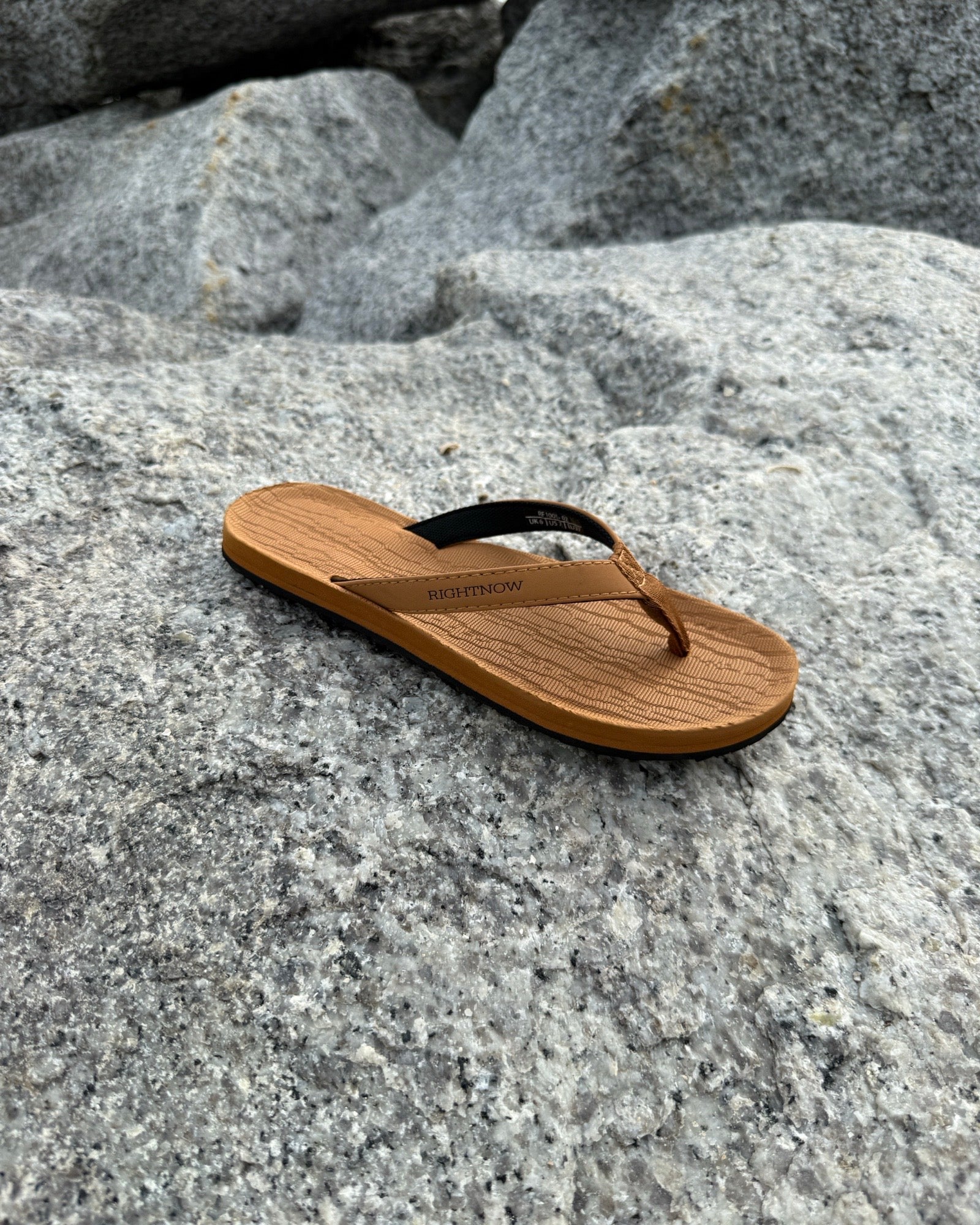 A single women's earth brown Flow Thong Sandal resting on rugged coastal rock, showcasing its minimalist design for effortless outdoor presence.