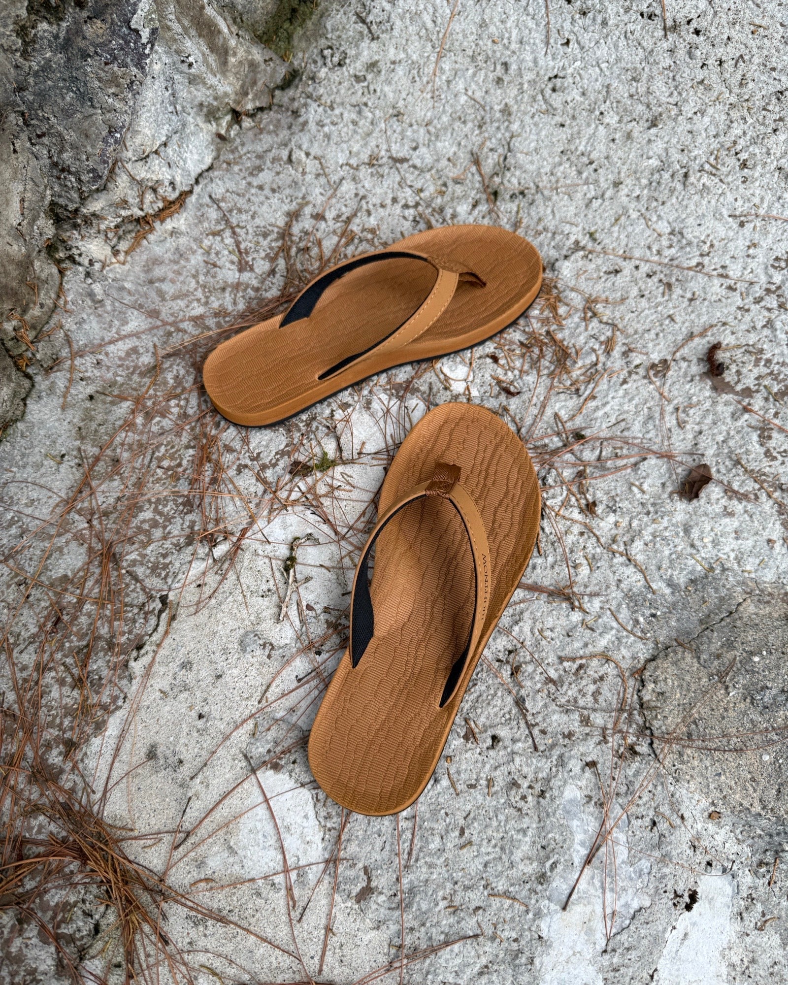 Top-down view of women's earth-tone Flow Thong Sandals on concrete ground with scattered pine needles, highlighting their simple design for natural and urban exploration.