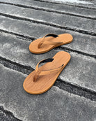 A pair of women's earth brown Flow Thong Sandals on a dark, textured concrete surface, showcasing their minimalist comfort for modern living.