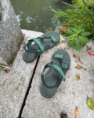 A pair of women's natural green EXPLOre Roman Sandals on a stone ledge by a tranquil stream, showing a simple, authentic lifestyle connected with nature.