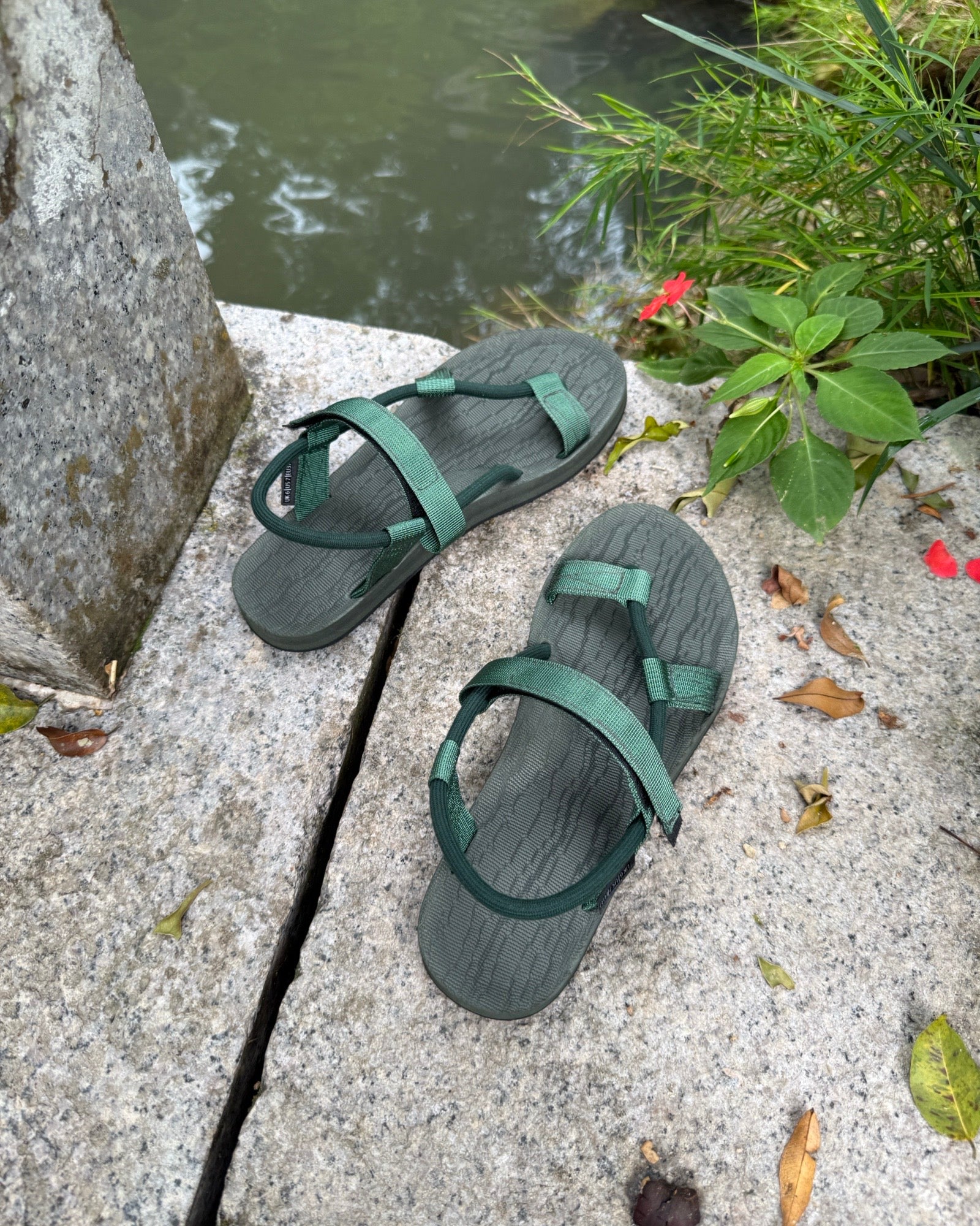 A pair of women's natural green EXPLOre Roman Sandals on a stone ledge by a tranquil stream, showing a simple, authentic lifestyle connected with nature.