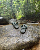 Women's natural green EXPLOre Roman Sandals on a river rock with a lush tropical rainforest backdrop, the perfect footwear for authentic tropical exploration.