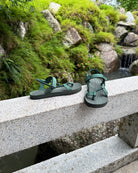 Women's natural green EXPLOre Roman Sandals on a bridge over a serene waterfall and lush greenery, perfect for travel and finding a quiet moment of presence in nature.