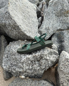 A single women's natural green EXPLOre Roman Sandal on a rugged coastal granite boulder, highlighting its durable minimalist strap design for all-terrain wear.