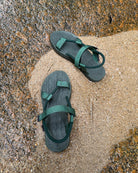 Top-down view of women's forest-tone EXPLOre Roman Sandals on wet, sandy riverside ground, showing their simple design and flexible straps for any natural terrain.