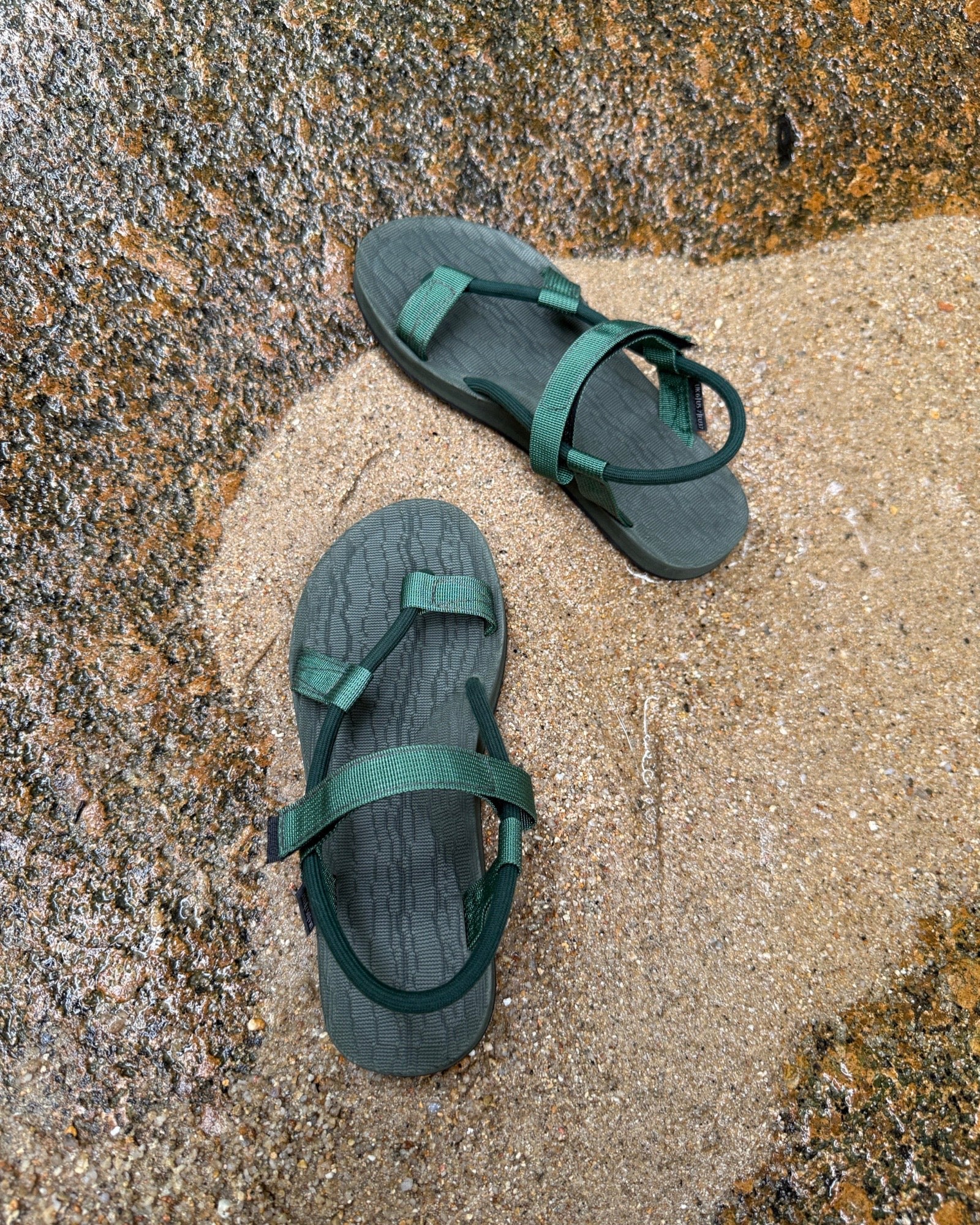 Top-down view of women's forest-tone EXPLOre Roman Sandals on wet, sandy riverside ground, showing their simple design and flexible straps for any natural terrain.