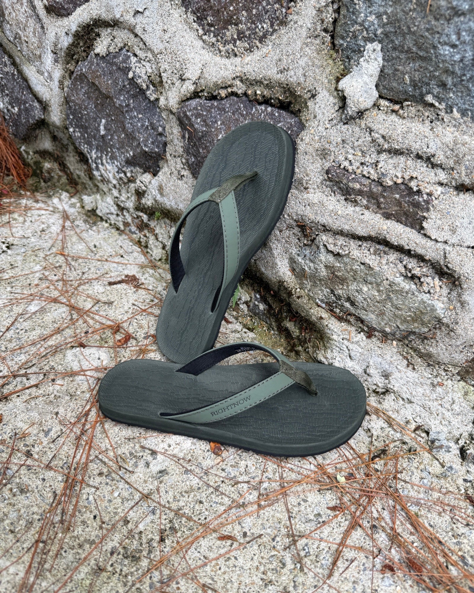 Women's natural green Flow Thong Sandals leaning against a textured stone wall, conveying a casual yet authentic style ready for everyday adventures.