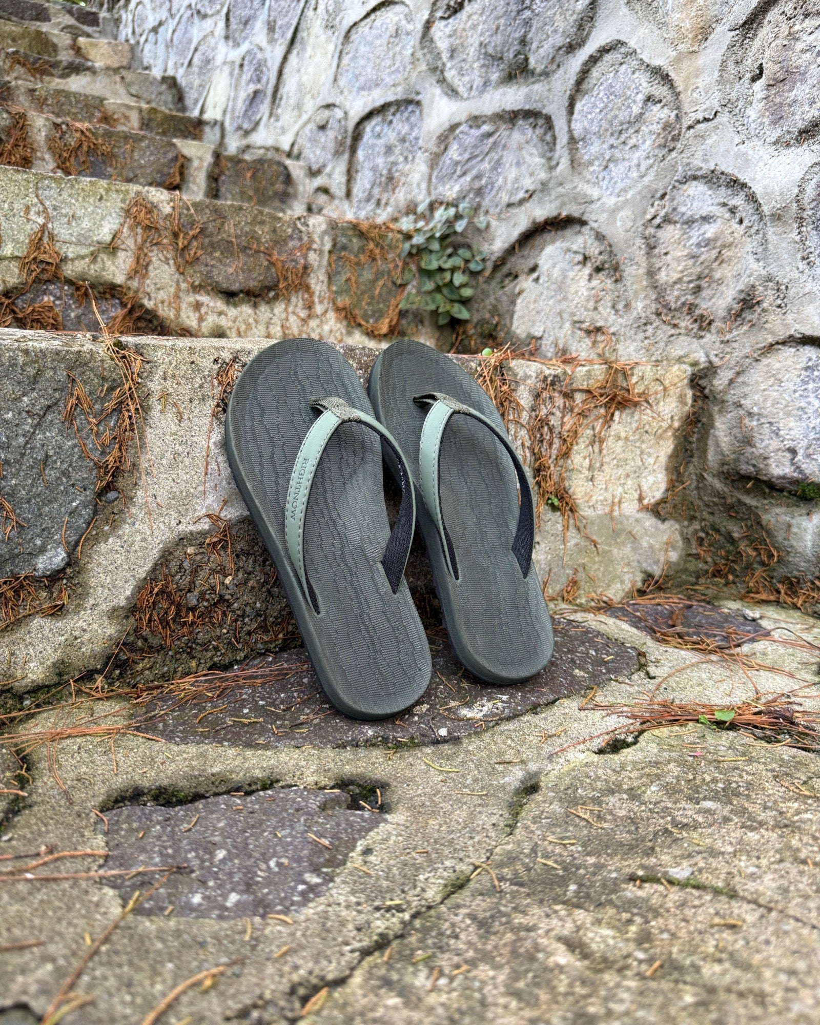A pair of women's natural green Flow Thong Sandals on rustic stone steps, perfect for a simple and authentic journey through historical or natural paths.