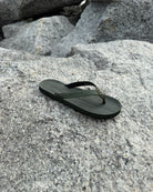 A single women's natural green Flow Thong Sandal resting on rugged coastal rock, showcasing its minimalist design for effortless outdoor presence.
