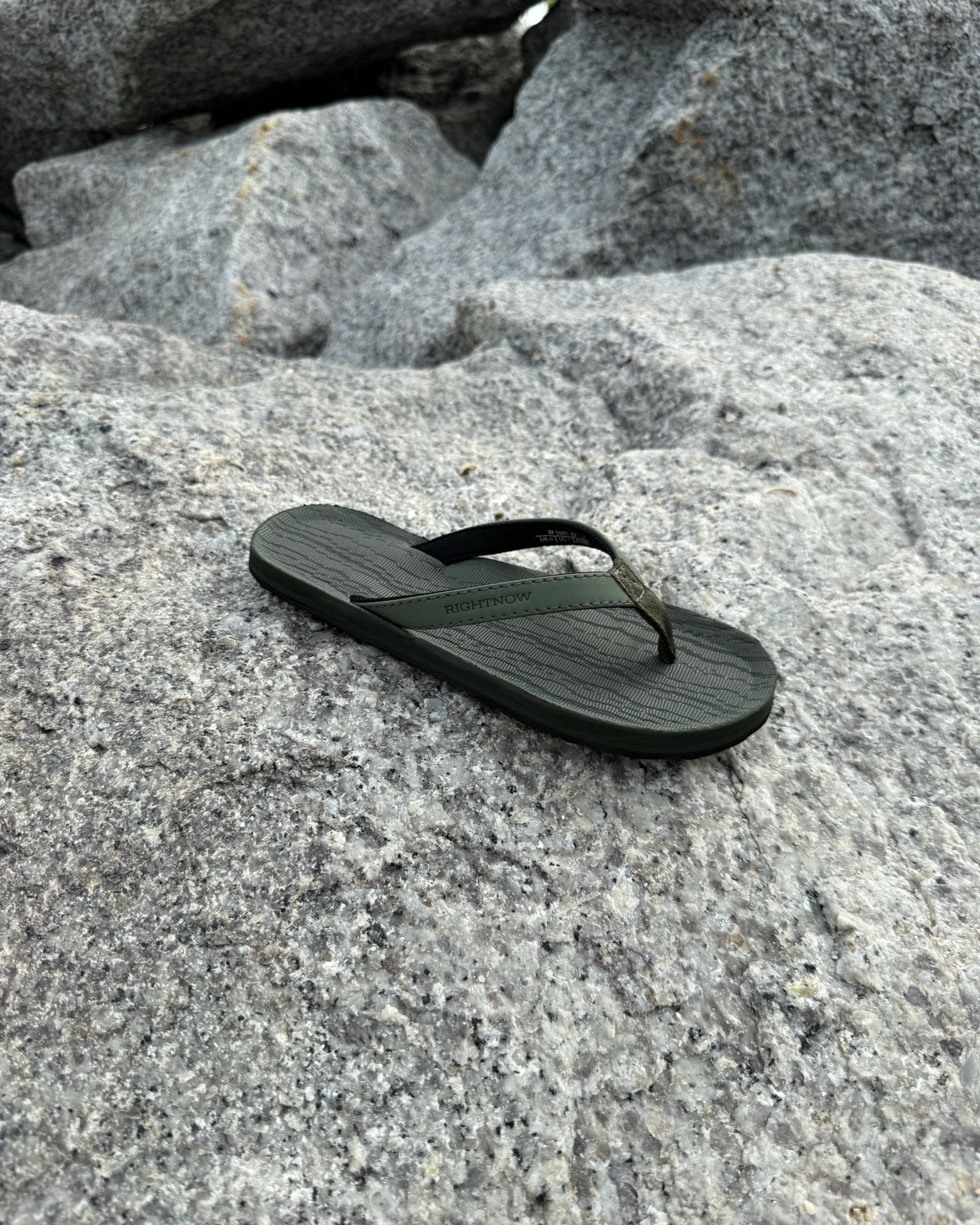 A single women's natural green Flow Thong Sandal resting on rugged coastal rock, showcasing its minimalist design for effortless outdoor presence.
