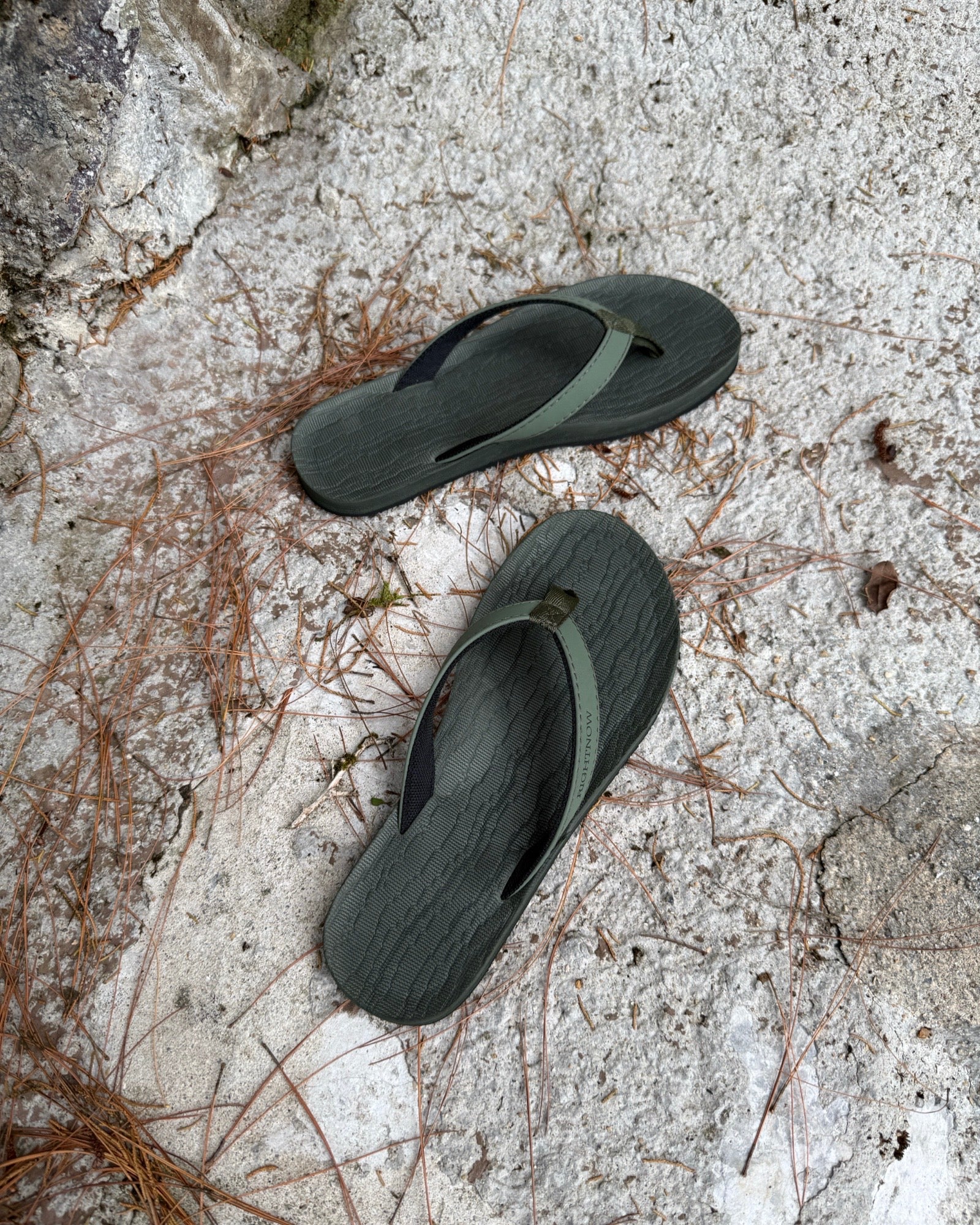 Top-down view of women's forest-tone Flow Thong Sandals on concrete ground with scattered pine needles, highlighting their simple design for natural and urban exploration.