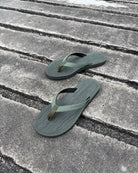 A pair of women's natural green Flow Thong Sandals on a dark, textured concrete surface, showcasing their minimalist comfort for modern living.