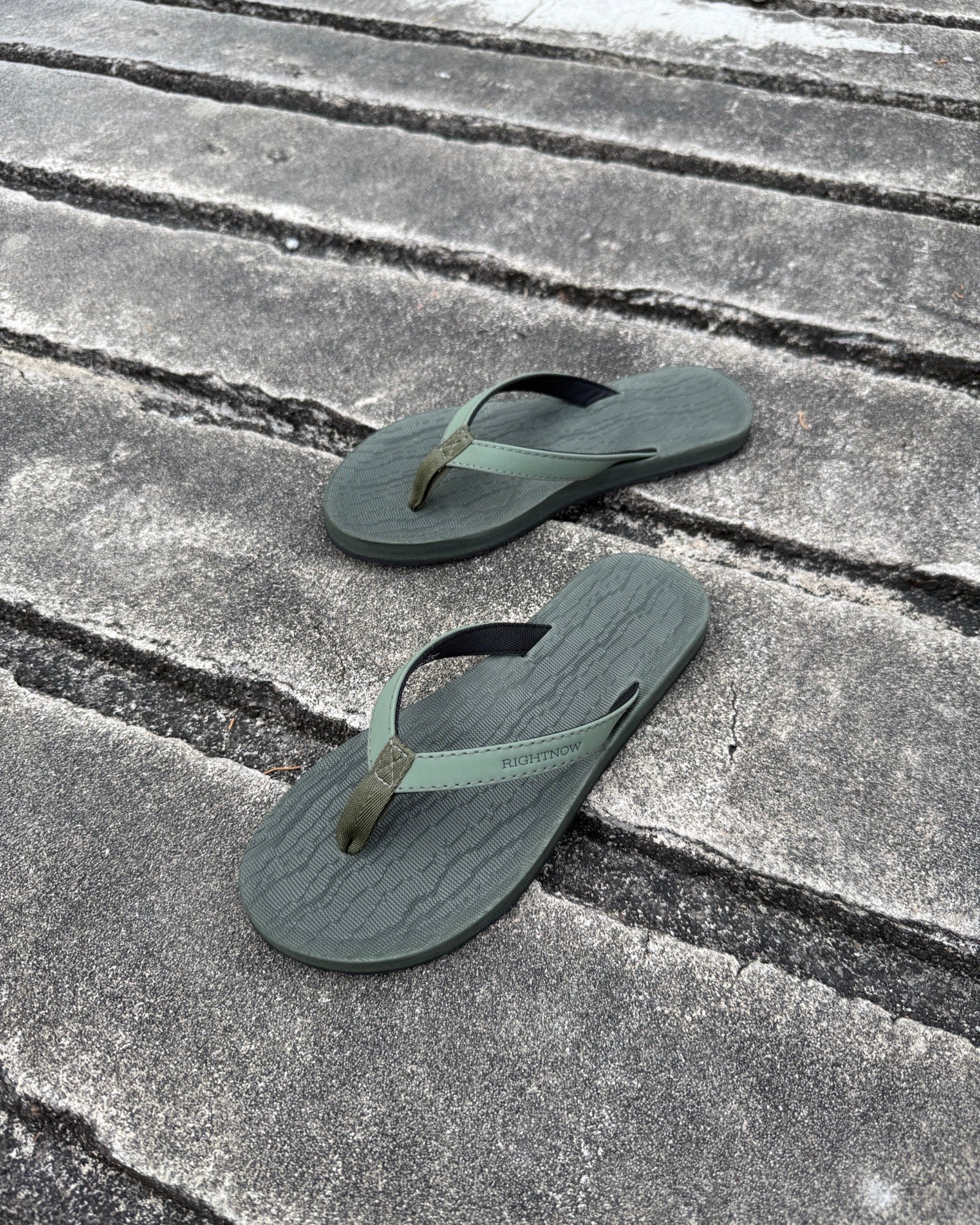 A pair of women's natural green Flow Thong Sandals on a dark, textured concrete surface, showcasing their minimalist comfort for modern living.
